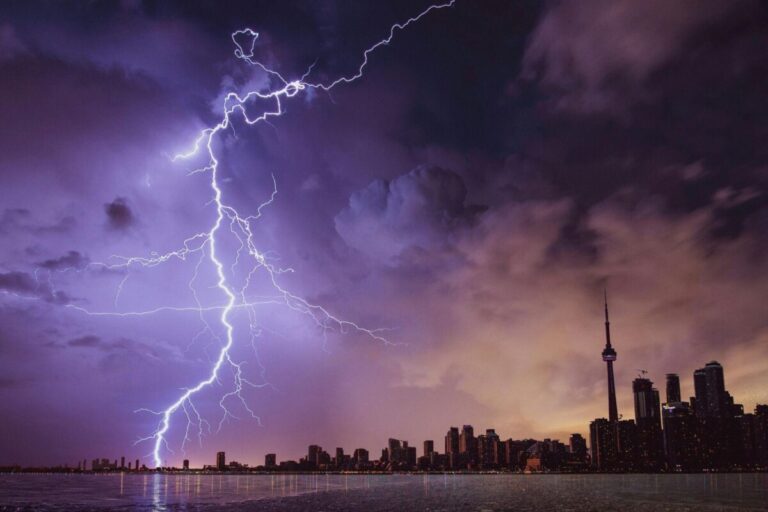 A large lightning bolt strikes above a city skyline at dusk, with dark clouds and the CN Tower visible in the background across a body of water.